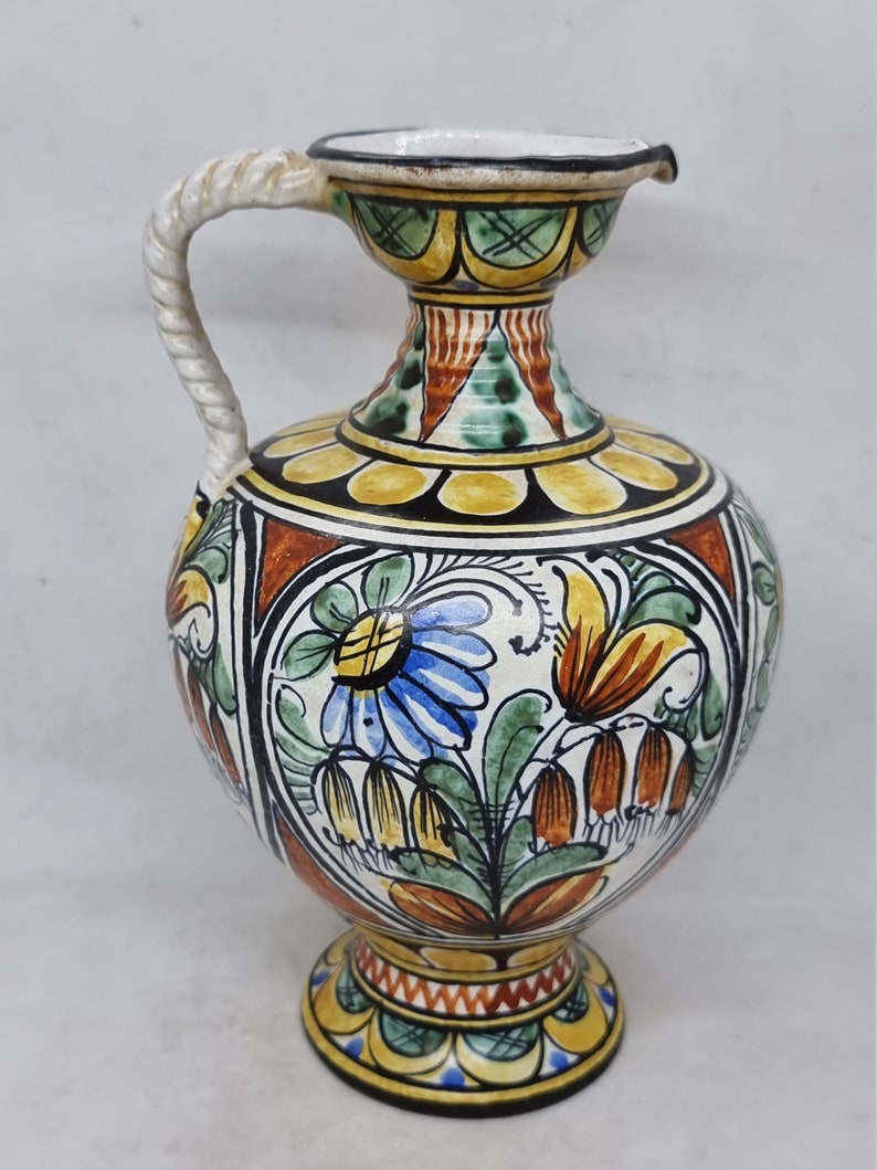 May include: A hand-painted ceramic pitcher with a white rope handle. The pitcher has a yellow, green, blue, and orange floral design.