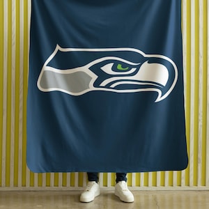 Puede incluir: Una manta azul marino con el logotipo de los Seattle Seahawks. El logotipo es una cabeza de águila estilizada en blanco, gris y verde. La manta se sostiene contra una pared a rayas amarillas y blancas.