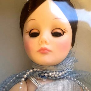 May include: A porcelain doll with brown hair, wearing a blue and silver dress with a pearl necklace. The doll has a crown on her head and is looking straight ahead.