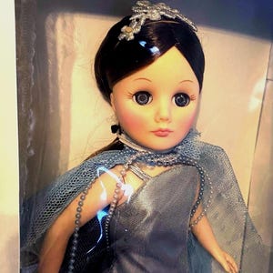 May include: A porcelain doll with dark brown hair, wearing a silver and blue dress with a pearl necklace. The doll has blue eyes and a silver crown.