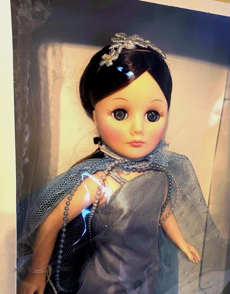 May include: A porcelain doll with dark brown hair, wearing a silver and blue dress with a pearl necklace. The doll has blue eyes and a silver crown.