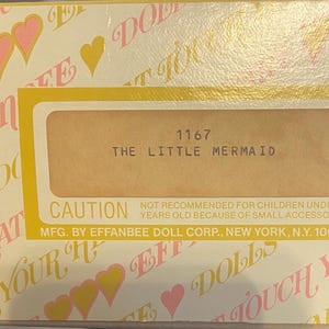 May include: A yellow and white box with a pink heart pattern. The box has a label that reads "1167 The Little Mermaid" and a caution label that reads "Not recommended for children under three years old because of small accessories. Mfg. by Effanbee Doll Corp., New York, N.Y. 10010, U.S.A."