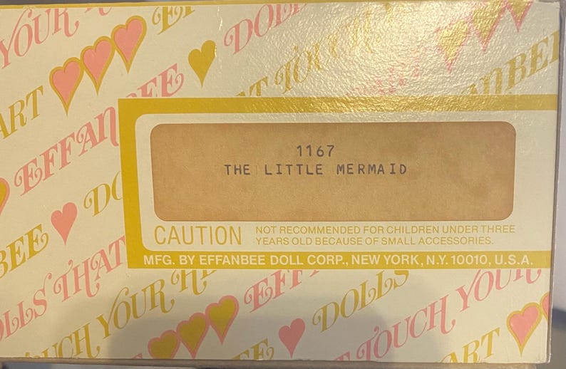 May include: A yellow and white box with a pink heart pattern. The box has a label that reads "1167 The Little Mermaid" and a caution label that reads "Not recommended for children under three years old because of small accessories. Mfg. by Effanbee Doll Corp., New York, N.Y. 10010, U.S.A."