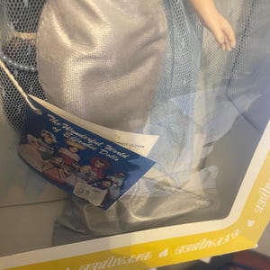 May include: A silver and blue doll wearing a long, flowing gown with a tulle overlay. The doll is part of "The Wonderful World of Eibabie Dolls" collection. The doll is packaged in a yellow box with a heart design.