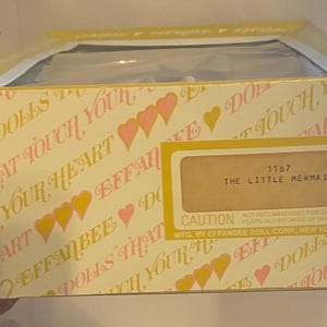 May include: A yellow and pink box with the words "Effanbee Dolls That Touch Your Heart" and "The Little Mermaid" printed on it. The box has a caution label and the number 1167 printed on it.