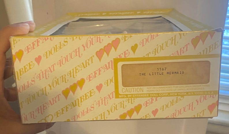 May include: A yellow and pink box with the words "Effanbee Dolls That Touch Your Heart" and "The Little Mermaid" printed on it. The box has a caution label and the number 1167 printed on it.