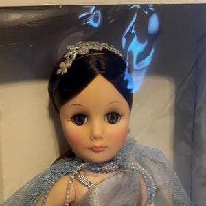 May include: A porcelain doll with brown hair, wearing a silver dress and a blue tulle cape. The doll has a silver headband with a floral design and large brown eyes.