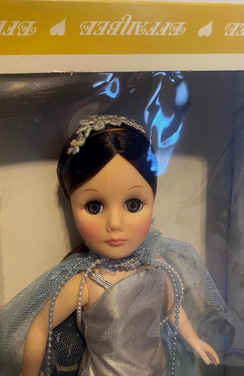 May include: A porcelain doll with brown hair, wearing a silver dress and a blue tulle cape. The doll has a silver headband with a floral design and large brown eyes.