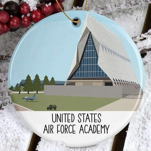 May include: A round ceramic ornament featuring a detailed illustration of the United States Air Force Academy. The design includes the academy's iconic chapel, a blue sky, and the text "UNITED STATES AIR FORCE ACADEMY".