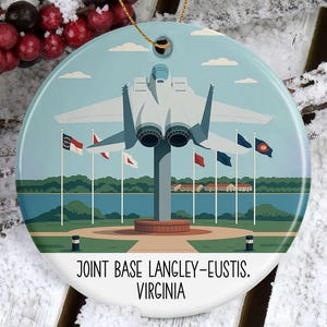 May include: Round ceramic ornament featuring a stylized illustration of an aircraft over a body of water with flags. The ornament has the text "JOINT BASE LANGLEY-EUSTIS. VIRGINIA" at the bottom. A gold string is attached for hanging.