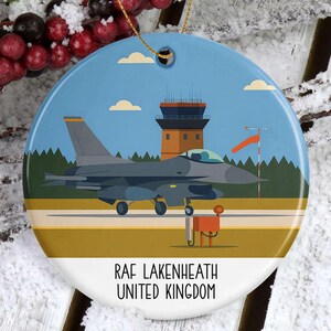 May include: A round ceramic ornament featuring an illustration of an F-16 fighter jet on a runway with a control tower in the background. The ornament has a blue sky and the text "RAF LAKENHEATH UNITED KINGDOM".