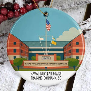 May include: A round ceramic ornament featuring a detailed illustration of the Naval Nuclear Power Training Command building. The ornament has a blue sky background with an American flag and the text "NAVAL NUCLEAR POWER TRAINING COMMAND, SC".