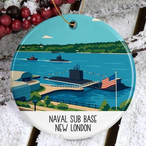 May include: A round ceramic ornament featuring a nautical scene with submarines, the American flag, and the text "Naval Sub Base New London." The ornament has a gold string for hanging and is set against a snowy, wooden background.