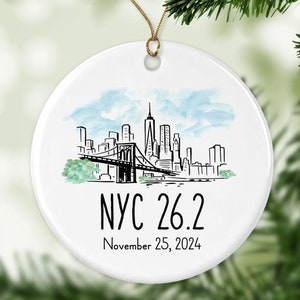 May include: A white ceramic Christmas ornament with a New York City skyline design. The ornament features a black and white illustration of the city skyline, the Brooklyn Bridge, and the text "NYC 26.2" and "November 25, 2024". The ornament is hanging from a gold string.