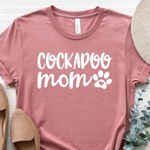 May include: A dusty rose t-shirt with the words "COCKAPOO mom" in white script, accompanied by a paw print graphic. The shirt is styled with tan loafers, a beige hat, and sprigs of greenery.