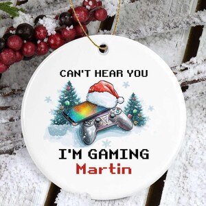 May include: White ceramic Christmas ornament with a gaming theme. The ornament features a controller, smartphone, and Santa hat design with the text "CAN'T HEAR YOU I'M GAMING Martin". Includes a gold string for hanging.