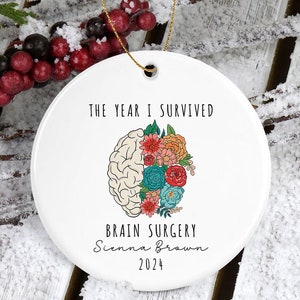 May include: White round ceramic ornament with the text "The year I survived brain surgery Sienna Brown 2024". A stylized brain with colorful flowers is depicted on the ornament.