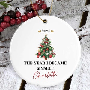 May include: White ceramic Christmas ornament with a gold string. The ornament features a decorated Christmas tree with red and green ornaments, the year "2021", and the text "THE YEAR I BECAME MYSELF" with the name "Charlotte" in red.