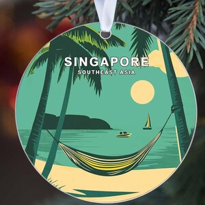 May include: Round Christmas ornament featuring a tropical beach scene with palm trees, a hammock, and sailboats. The ornament has the word "SINGAPORE" and "SOUTHEAST ASIA" printed on it. The background is a teal color with a yellow sun.