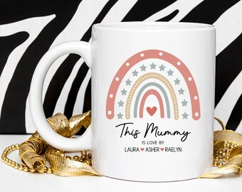 May include: White ceramic mug featuring a rainbow design in shades of pink, beige, and blue, with star accents. The text "This Mummy is loved by Laura, Asher, Raelyn" is printed below the rainbow. A gold beaded necklace is in the foreground.