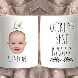 May include: Two white ceramic mugs with black text and a baby's face on one mug. The mug on the left says "Love Weston" and has a baby's face. The mug on the right says "World's Best Nanny".