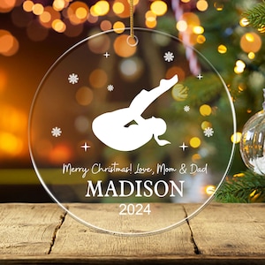 May include: Clear, round Christmas ornament with a gymnast silhouette and snowflakes. The ornament has the text "Merry Christmas! Love, Mom & Dad" and the name "MADISON" with the year "2024". The ornament hangs from a gold string.