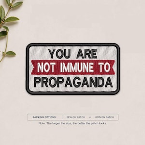 May include: A rectangular embroidered patch with a white background and a black border. The text reads YOU ARE in black, NOT IMMUNE TO in white on a red banner, and PROPAGANDA in black. Below, text states BACKING OPTIONS: SEW ON PATCH or IRON ON PATCH.
