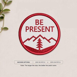 May include: A circular embroidered patch featuring a white background with a red border. The text BE PRESENT is stitched in red above a red mountain range and a single pine tree. Below the patch, text reads BACKING OPTIONS: SEW ON PATCH or IRON ON PATCH.
