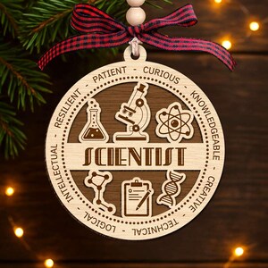 May include: A wooden Christmas ornament with the word "SCIENTIST" in the center. The ornament features science-related icons and words like "Patient" and "Curious" around the edge. It is decorated with a red and black plaid ribbon.
