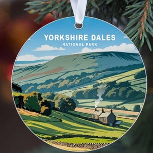May include: Round Christmas ornament featuring a landscape illustration of the Yorkshire Dales National Park. The design includes rolling green hills, a small cottage, and the text "Yorkshire Dales National Park" in white lettering against a blue sky.
