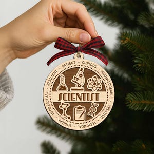 May include: A round wooden ornament with the word "SCIENTIST" and science-related icons. The ornament has a dark brown center and a light brown outer ring with words like "Patient" and "Curious". It is held by a hand and has a red and black plaid ribbon.