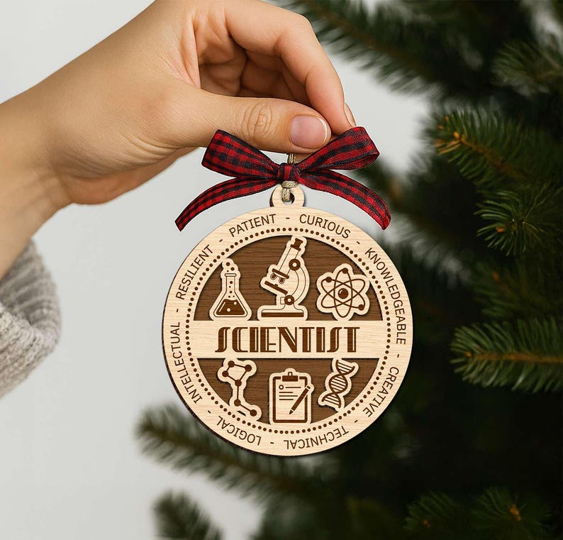 May include: A round wooden ornament with the word "SCIENTIST" and science-related icons. The ornament has a dark brown center and a light brown outer ring with words like "Patient" and "Curious". It is held by a hand and has a red and black plaid ribbon.