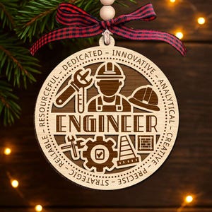 May include: A wooden Christmas ornament with the word "ENGINEER" in the center. The ornament features an engineer graphic, tools, and the words "RESOURCEFUL, RELIABLE, STRATEGIC, PRECISE, CREATIVE, ANALYTICAL, INNOVATIVE, DEDICATED". A red and black plaid ribbon is attached.