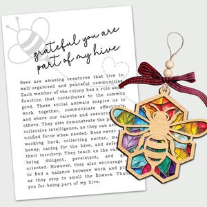 May include: Wooden bee ornament with a stained glass design in a honeycomb shape, accented with a red ribbon and wooden beads. The ornament hangs next to a card with the text "grateful you are part of my hive."