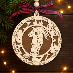 May include: Wooden golf-themed Christmas ornament. The ornament features a golfer in the center, surrounded by golf terms like "Fairway" and "Birdie." It is adorned with a red and black plaid ribbon and wooden beads for hanging.