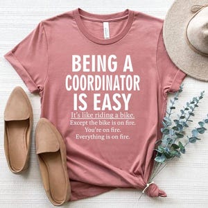 May include: A pink t-shirt with the text "BEING A COORDINATOR IS EASY. It's like riding a bike. Except the bike is on fire. You're on fire. Everything is on fire."