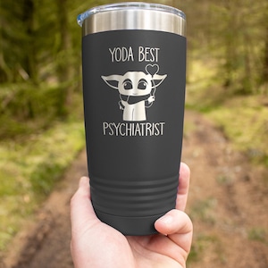 Puede incluir: Un vaso de acero inoxidable gris oscuro con un dise&ntilde;o blanco de un personaje de dibujos animados sosteniendo un globo en forma de coraz&oacute;n. El texto "YODA BEST PSYCHIATRIST" est&aacute; impreso en el vaso.