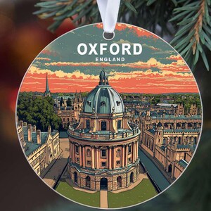 May include: Round Christmas ornament featuring a detailed illustration of Oxford, England, with the text "OXFORD ENGLAND" at the top. The image showcases a prominent building with a dome, set against a colorful sky. A white ribbon is attached for hanging.