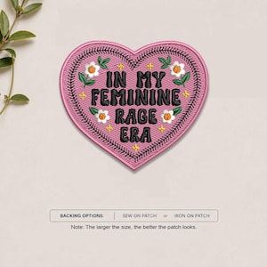 May include: A heart-shaped embroidered patch in pink with black text that reads IN MY FEMININE RAGE ERA. The patch features white flowers with yellow centers, green leaves, and small yellow star accents, finished with a black stitched border.