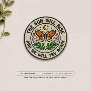 May include: A circular embroidered patch featuring an orange butterfly, pink flowers, green leaves, and a crescent moon. The text reads THE SUN WILL RISE AND WE WILL TRY AGAIN. Below, text states BACKING OPTIONS: SEW ON PATCH or IRON ON PATCH.
