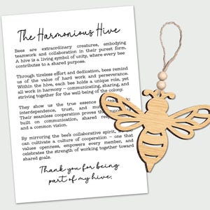 May include: A wooden bee-shaped ornament with a string for hanging, next to a white paper with the text "The Harmonious Hive". The bee has cut-out details and a natural wood grain finish. The paper describes the value of teamwork.
