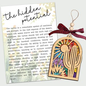 May include: Wooden cactus ornament with a stained glass design featuring a sun and desert plants. The ornament is tied with a red and black ribbon and has the text "The hidden potential" on a card.