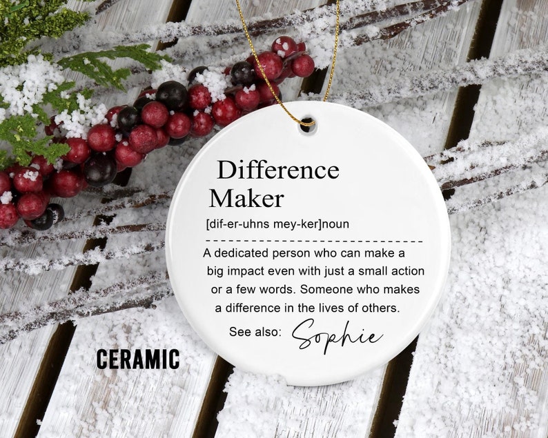 May include: A white ceramic ornament with the text "Difference Maker" and a definition. The ornament has a gold string for hanging. The word "CERAMIC" is printed at the bottom. The background includes snowy branches and berries.