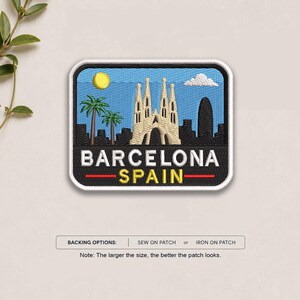 May include: A rectangular embroidered patch featuring a blue sky with a yellow sun and white cloud above a black silhouette of the Barcelona skyline, including the Sagrada Familia. The text reads BARCELONA SPAIN in white and yellow letters.