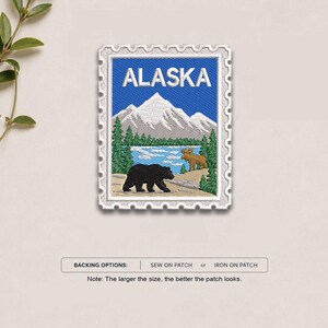 May include: A rectangular embroidered patch shaped like a postage stamp featuring the word ALASKA in white text. The design depicts a snow-capped mountain, a blue lake, green pine trees, a black bear, and a brown moose. Backing options include sew-on or iron-on.
