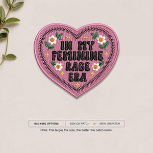 May include: A heart-shaped embroidered patch in pink with black text that reads IN MY FEMININE RAGE ERA. The design features small white flowers with yellow centers, green leaves, and tiny yellow stars. A black stitched border outlines the heart.