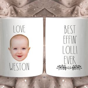 May include: Two white ceramic mugs with black text and a baby's face on one mug. The mug on the left says "Love Weston" and has a baby's face in the center. The mug on the right says "Best Effin' Lolli Ever" with a decorative leaf design at the bottom.