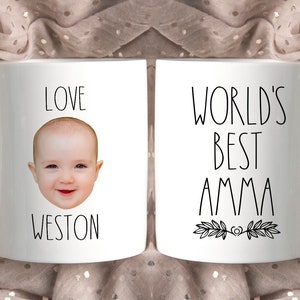 May include: Two white ceramic mugs with black text and a baby's face on one mug. The mug on the left says "Love Weston" and has a baby's face in the center. The mug on the right says "World's Best Amma" with a floral design at the bottom.