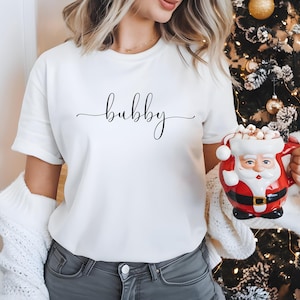 May include: White t-shirt with the word "bubbly" written in black cursive script. The shirt is being worn by a person holding a red Santa mug with marshmallows.