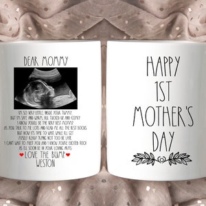 May include: White ceramic mug with a black and white ultrasound image and text that reads "Dear Mommy" and "Love the Bump Weston". The other side of the mug has black text that reads "Happy 1st Mother's Day" with a black floral design.